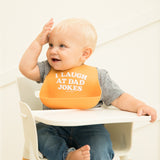 Babt wearing Bella Tunno I Laugh at Dad Jokes Wonder Bib in orange, featuring a "I Laugh at Dad Jokes" design, adjustable neck, and a catch pocket, made from BPA-free silicone, available at Be Charmed