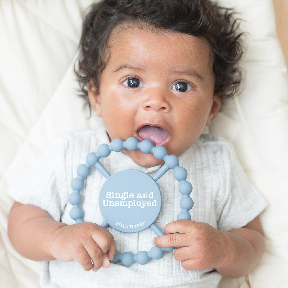 Baby holding Bella Tunno Single and Unemployed Teether in pastel blue, featuring a humorous "Single and Unemployed" message, made from BPA-free silicone, available at Be Charmed