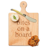 Wooden cutting board with 'bites on a board' engraving, knife, and food items on a white background