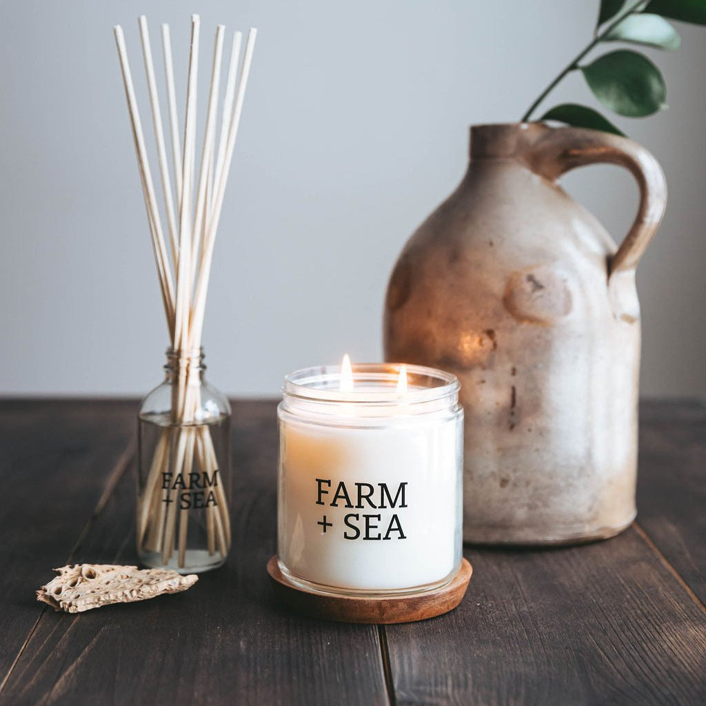 Farm + Sea  Peony + Sea Salt 14 oz Candle lit on table next to reed diffuser
