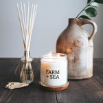 Farm + Sea  Peony + Sea Salt 14 oz Candle lit on table next to reed diffuser