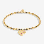 Gold bracelet with a heart-shaped tag featuring a pink gemstone on a white background. Available at Be Charmed