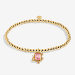 A gold beaded bracelet with a central gold-toned setting holding a rectangular pink gemstone, photographed on a clean white background. Available at Be Charmed