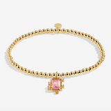 A gold beaded bracelet with a central gold-toned setting holding a rectangular pink gemstone, photographed on a clean white background. Available at Be Charmed