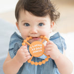Baby using a Bella Tunno "I Laugh at Dad Jokes" Teether in orange, featuring a beaded texture and easy-grip design, available at Be Charmed.
