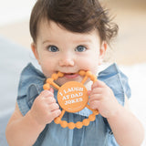 Baby using a Bella Tunno "I Laugh at Dad Jokes" Teether in orange, featuring a beaded texture and easy-grip design, available at Be Charmed.
