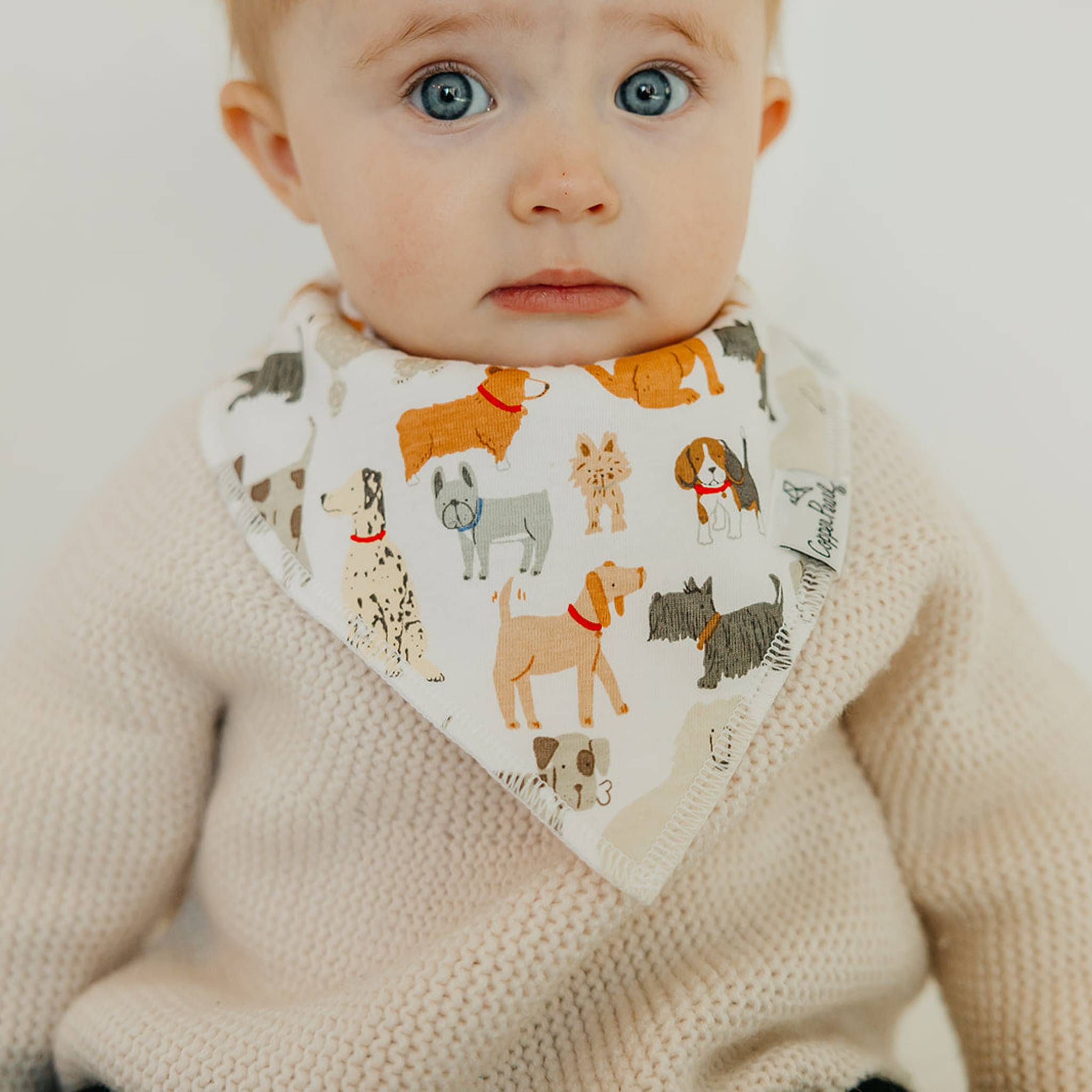Baby wearing a Copper Pearl Rufus Bandana Bib featuring absorbent cotton front and soft fleece back, adjustable for babies 0-36 months. Available at Be Charmed.