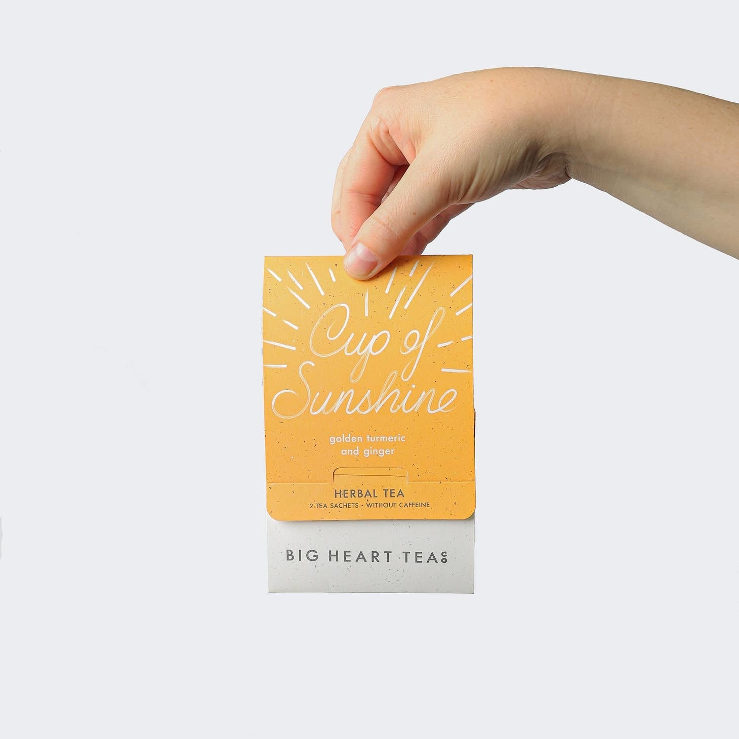 A hand is holding a tea bag with 'CUP OF SUNSHINE' printed on it, with the brand 'BIG HEART TEAS' visible below. The tea bag has a yellow color with a bright light background. Available at Be Charmed
