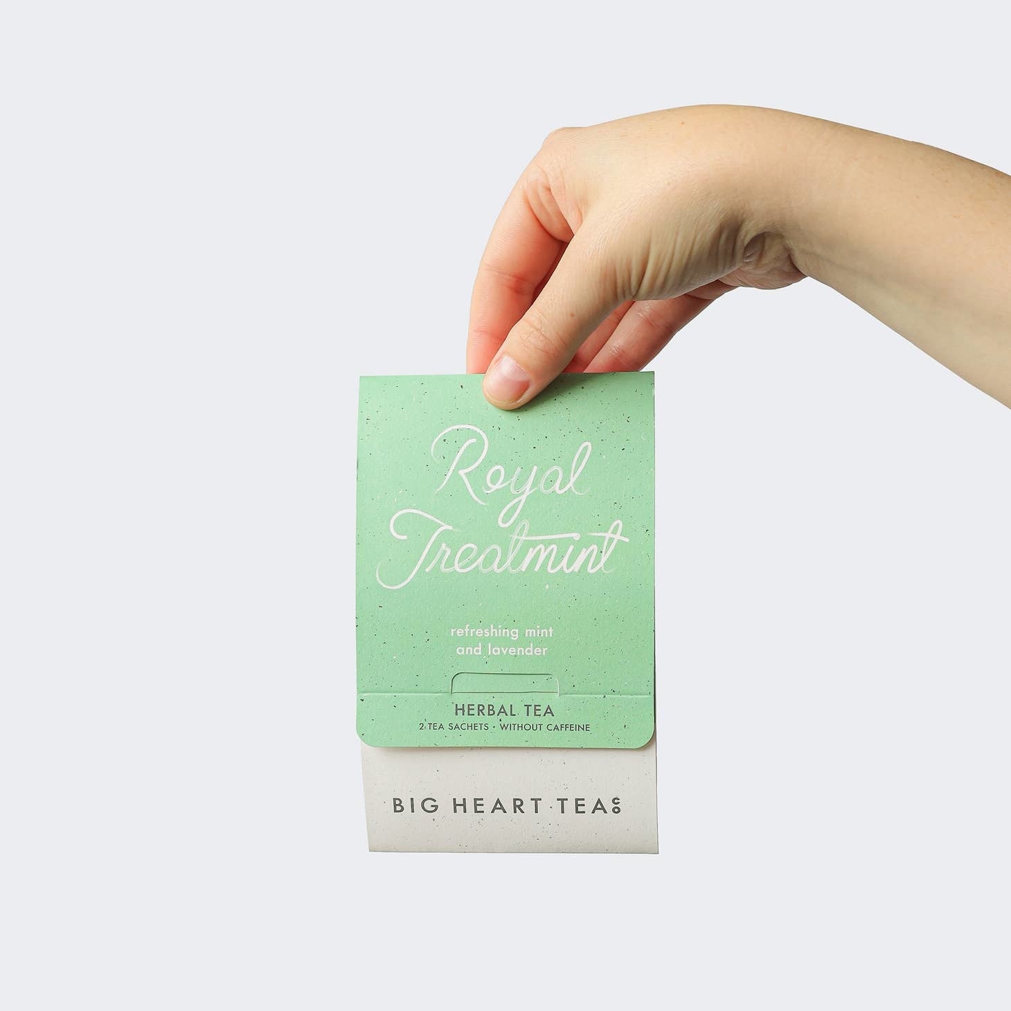 A hand holding a tea bag labeled 'Royal Treatment Mint' from Big Heart Tea Co. with organic tea leaves visible on the tag. Available at Be Charmed