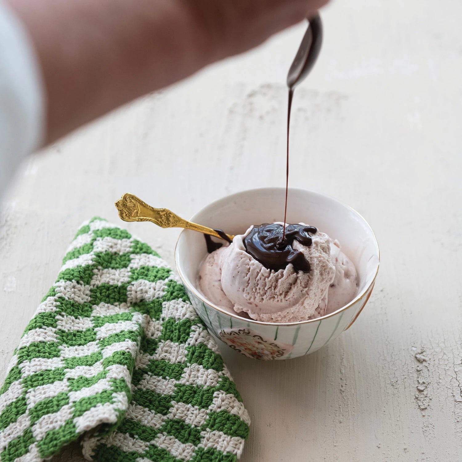Bowl of ice cream with chocolate sauce being drizzled over it on a light surface. Available at Be Charmed