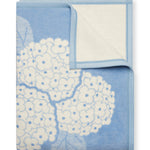 ChappyWrap Hydrangeas Bluebell Blanket featuring soft blue and lavender floral hydrangeas on a cream background. Made from a plush cotton blend for luxurious comfort and durability—ideal for garden-inspired décor or cozy lounging. Available at Be Charmed.
