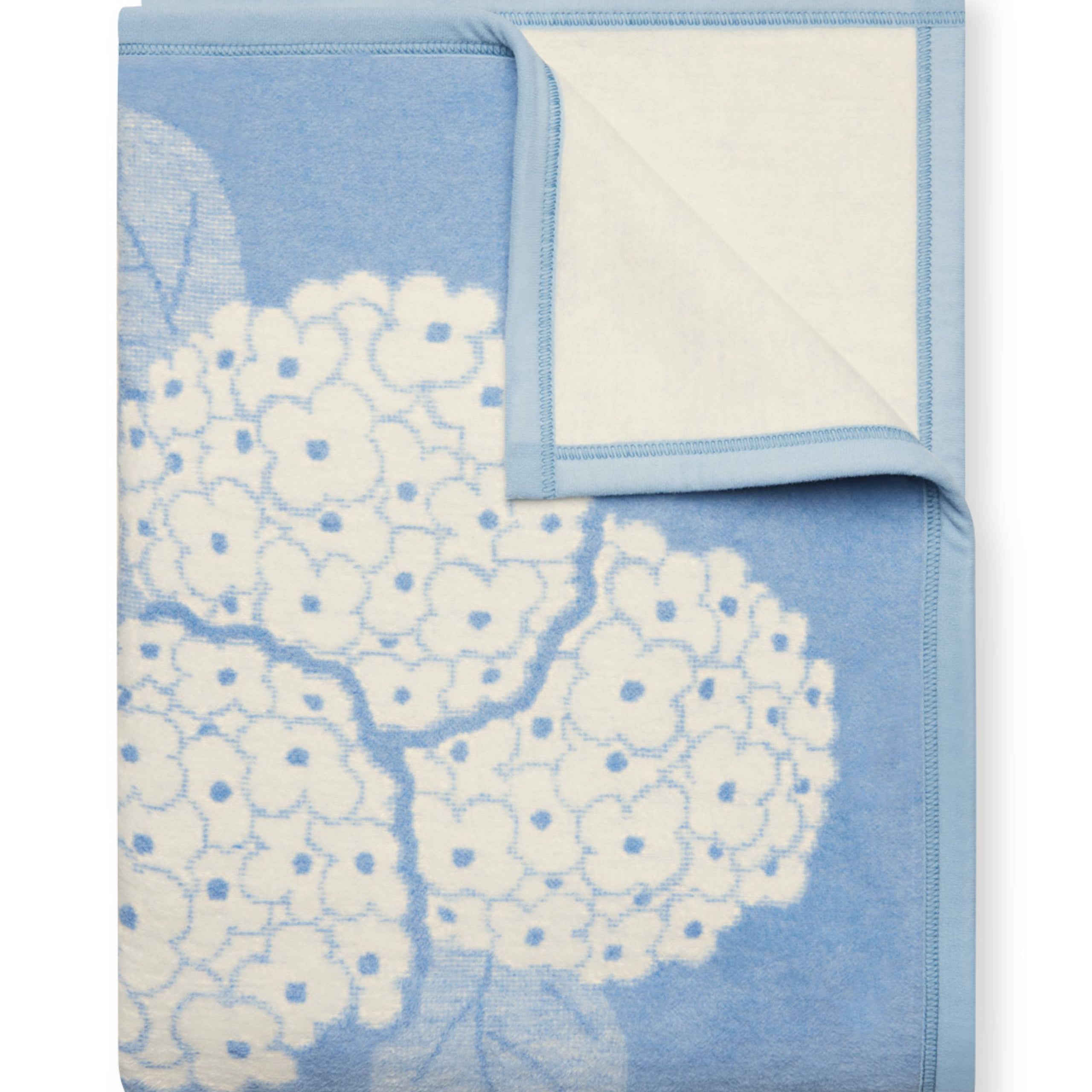ChappyWrap Hydrangeas Bluebell Blanket featuring soft blue and lavender floral hydrangeas on a cream background. Made from a plush cotton blend for luxurious comfort and durability—ideal for garden-inspired décor or cozy lounging. Available at Be Charmed.