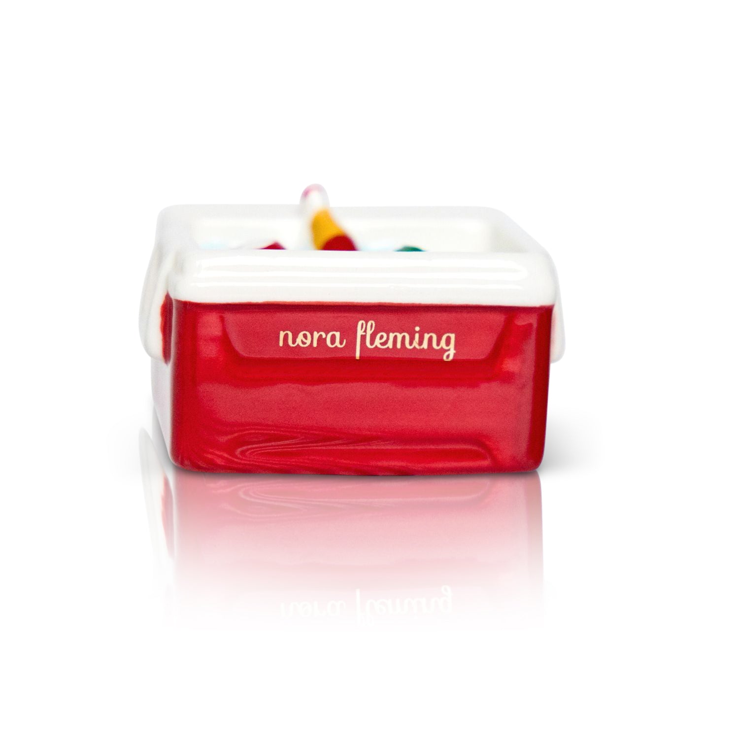 Red toy box with 'nora Fleming' branding on a white background. Available at Be Charmed