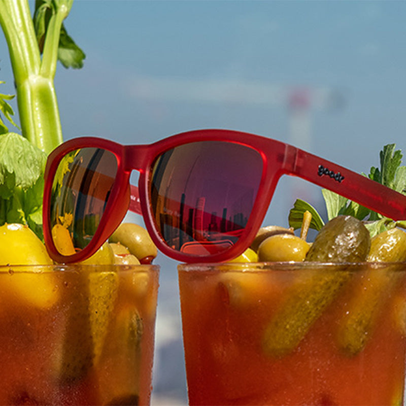Red Goodr sunglasses placed on top of a glass containing a bloody mary cocktail, garnished with a pickle and celery. Available at Be Charmed