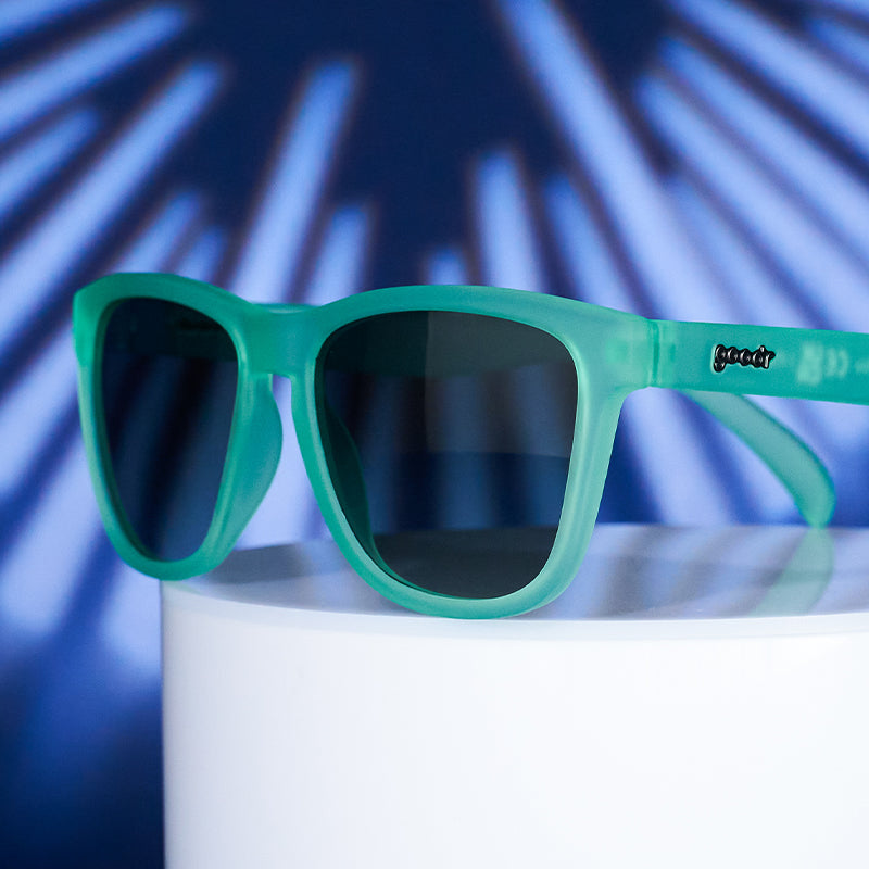 A pair of mint green sunglasses with dark green gradient lenses placed on a cylindrical stand with a blue and black background. Available at Be Charmed