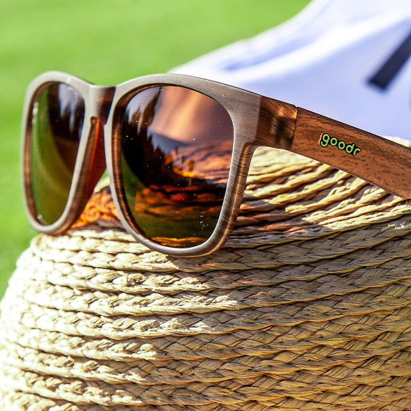A pair of dark-framed sunglasses with 'Goodr' branding on the side, resting on a straw hat. Available at Be Charmed