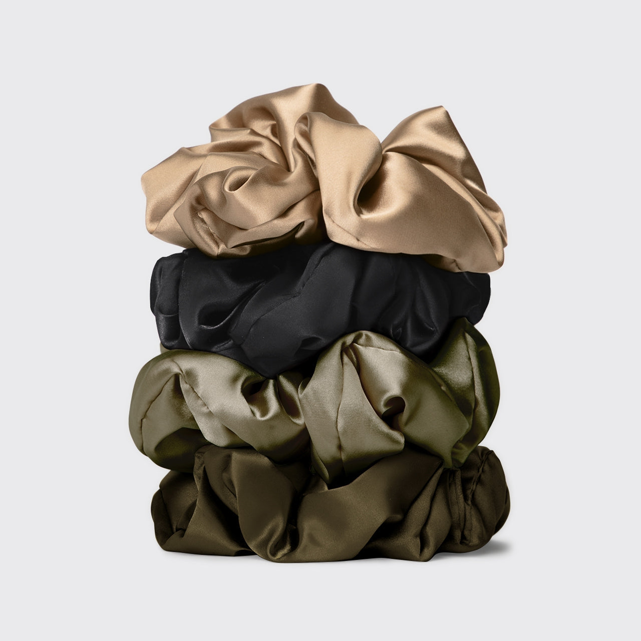 Stack of satin scrunchies in beige, black, and olive green on a light gray background. Available at Be Charmed