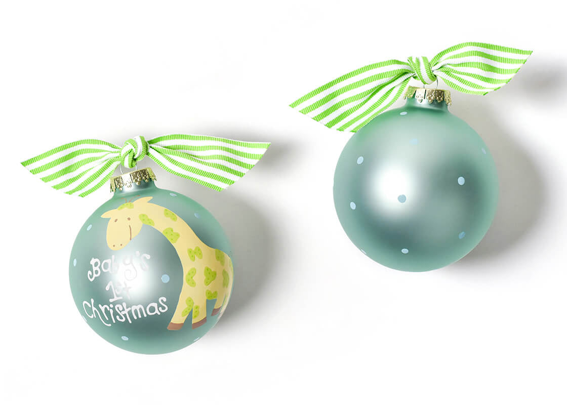 Two round glass ornaments with a Christmas theme. One has a green and white color scheme with a giraffe illustration and the text 'Baby's 1st Christmas'. Available at Be Charmed