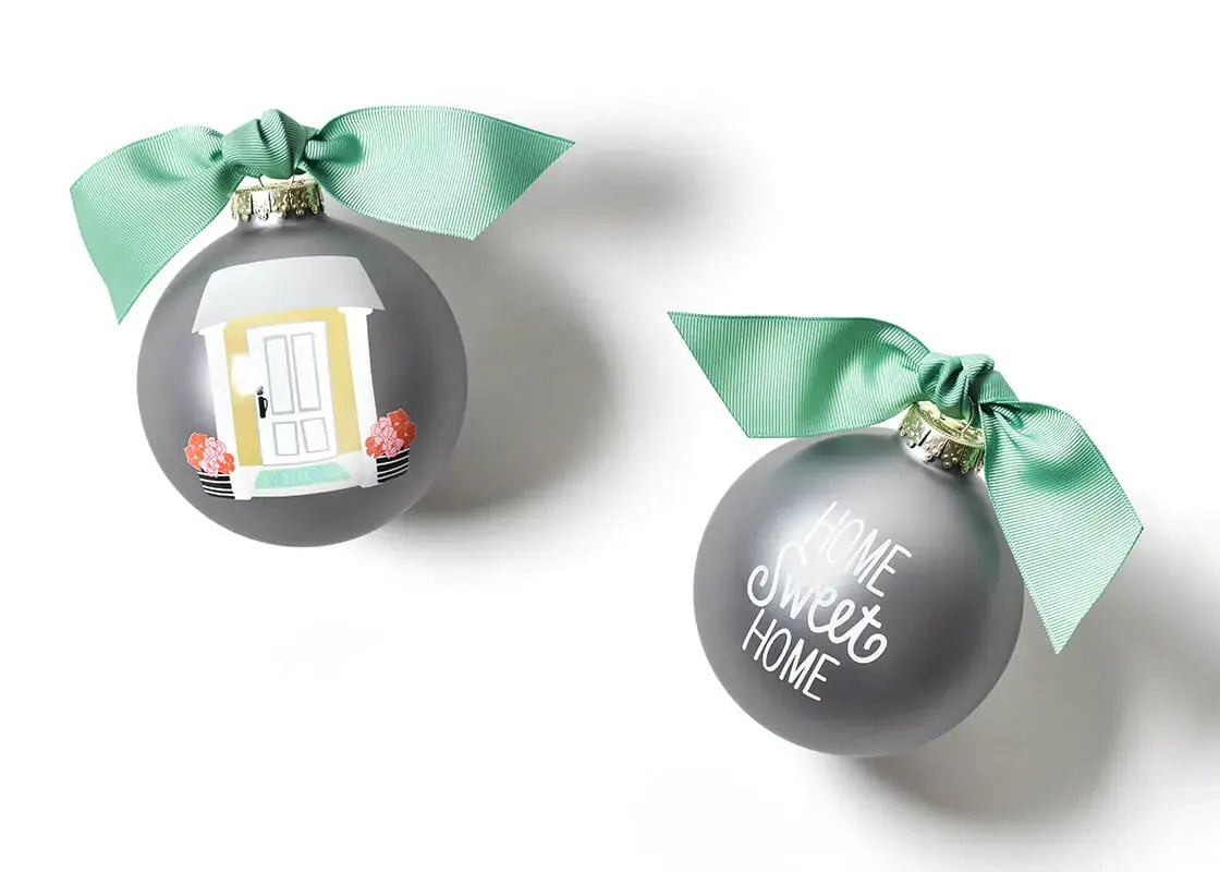 Two round glass ornaments with a hand-painted sketch of a front door on a silver background, white script, and a green accent ribbon on top. One ornament has a green bow on top. Available at Be Charmed