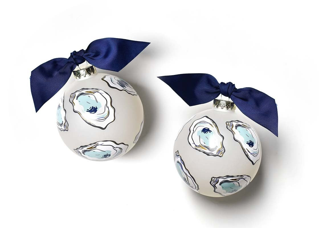 Two spherical glass ornaments with hand-drawn oysters in shades of blue and gold on a white background, adorned with navy blue bows. Available at Be Charmed