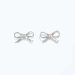 A pair of silver ribbon bow stud earrings on a white background. Available at Be Charmed