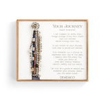 A gray-colored Demdaco bracelet with a heart pendant, featuring a series of seven beaded strands and a rope-like closure. The bracelet is presented in a box with a poetic message. Available at Be Charmed