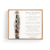 A gray-colored Demdaco bracelet with a heart pendant, featuring a series of seven beaded strands and a rope-like closure. The bracelet is presented in a box with a poetic message. Available at Be Charmed