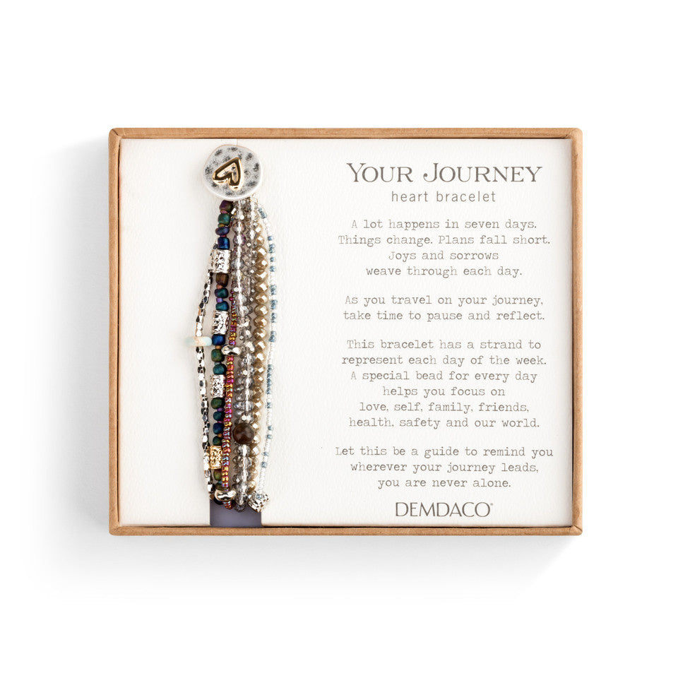 A gray-colored Demdaco bracelet with a heart pendant, featuring a series of seven beaded strands and a rope-like closure. The bracelet is presented in a box with a poetic message. Available at Be Charmed