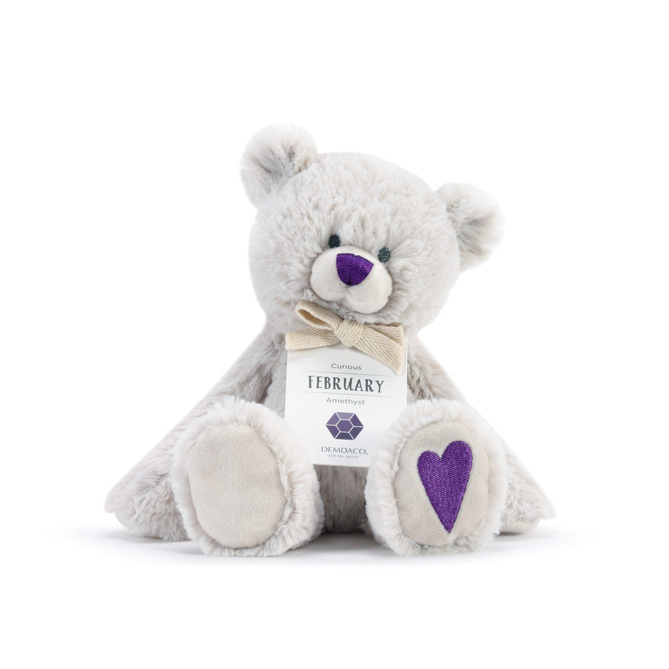 A plush teddy bear with a birthstone theme, featuring a ribbon around its neck that indicates the month of February. Available at Be Charmed.