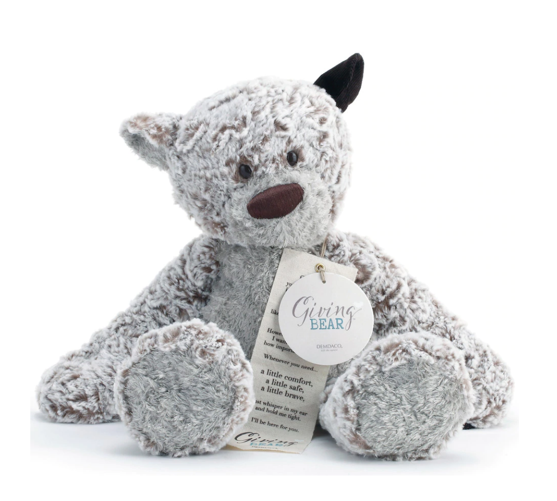 A plush teddy bear with weighted paws, extra-long arms, and a corduroy ear, sitting upright with a keepsake bookmark attached to its neck. Available at Be Charmed