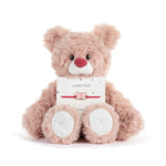 Plush teddy bear holding a 'Love You' card and bracelet on a white background. Availabl at Be Charmed