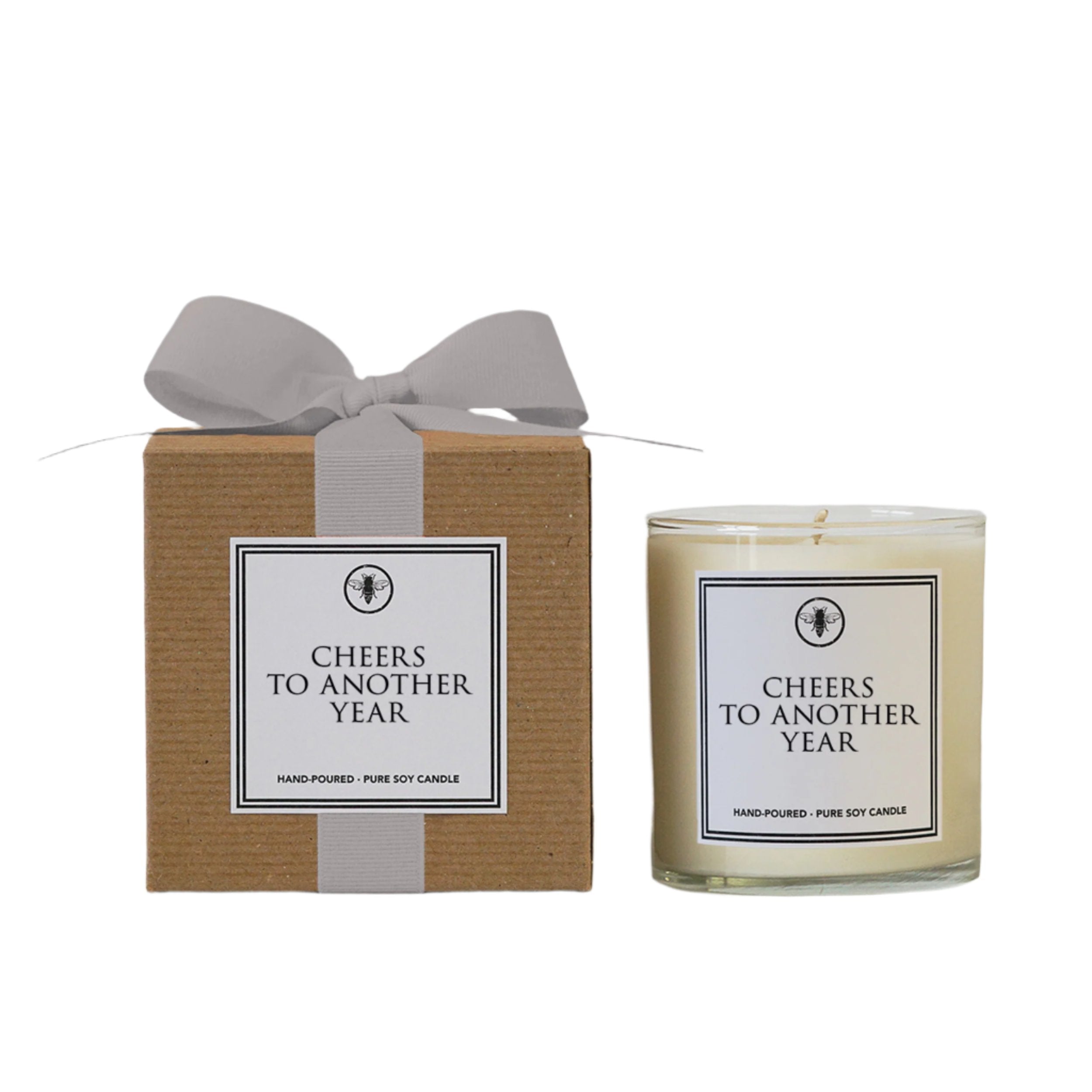 A beige candle with a label 'Cheers to another year' placed in a brown cardboard box with a grey ribbon bow on top, and a white label on the candle. Available at Be Charmed