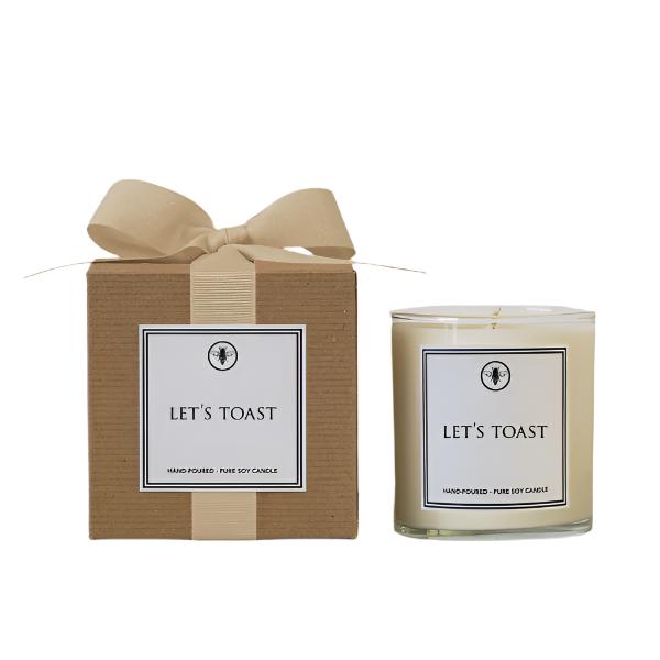 A beige candle with a label that reads 'Let's Toast' and a small bow on top, next to its cardboard packaging with the same text and a logo. Available at Be Charmed