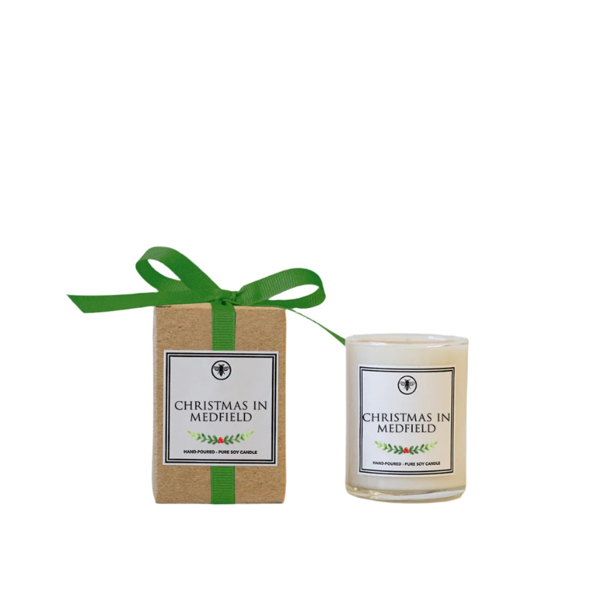 A candle with a label 'Christmas in Medfield' next to its recycled kraft box packaging adorned with a green grosgrain bow. Available at Be Charmed