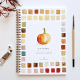A spiral-bound watercolor workbook titled 'AUTUMN' with a painted orange pumpkin in the center and a border of watercolor color swatches, shown on a white background with a paintbrush and paint tube nearby. Available at Be Charmed