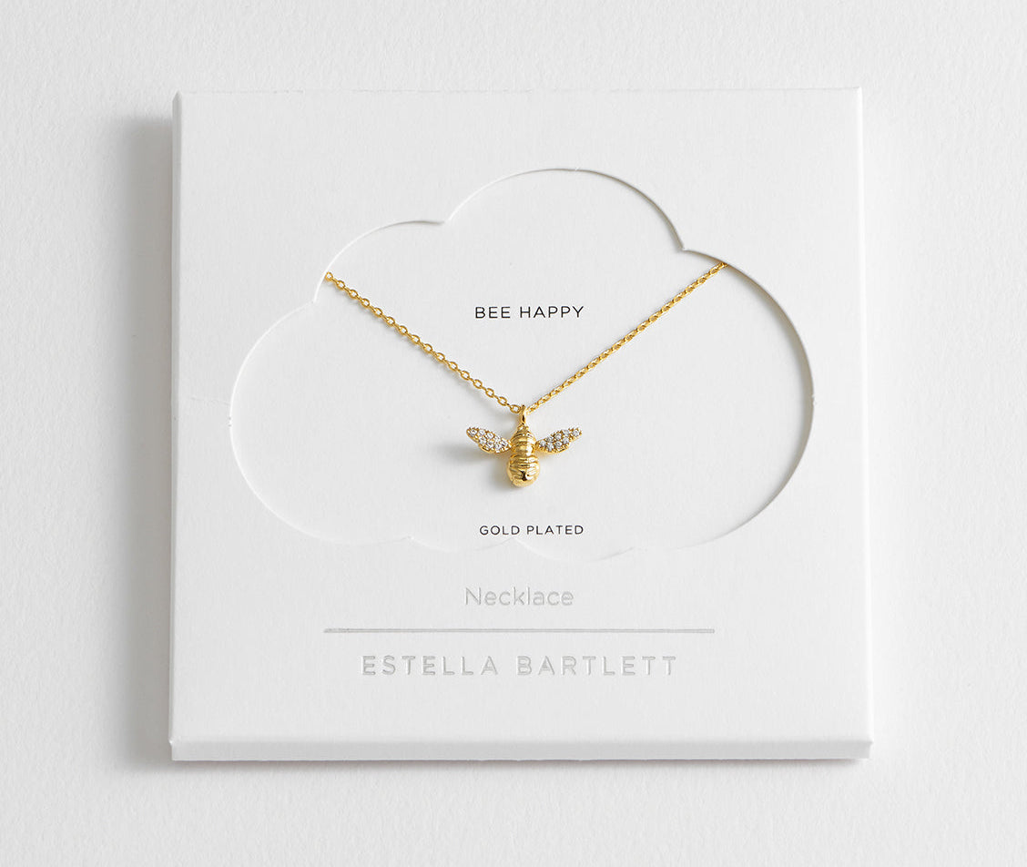 Gold bee necklace on a white card with 'Estella Bartlett' branding. Available at Be Charmed