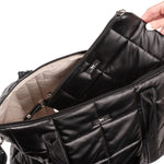 Black quilted Scout bag with a hand opening it on a light white background. Available at Be Charmed
