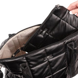 Black quilted Scout bag with a hand opening it on a light white background. Available at Be Charmed