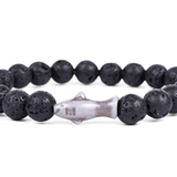 A beaded stretch Fahlo bracelet made of black porous lava stones with a central shark  charm, shown on a plain white background. Available at Be Charmed