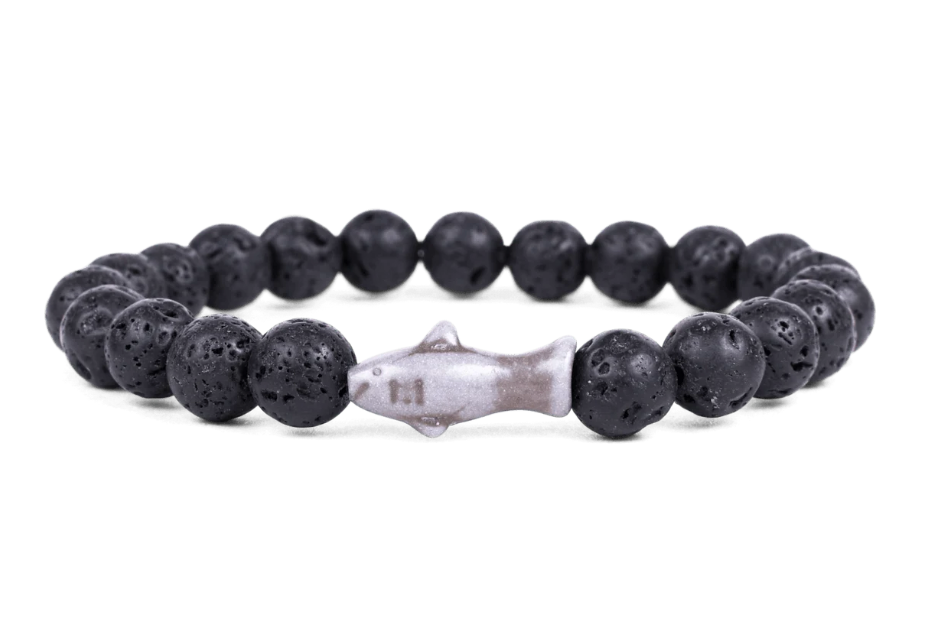 A beaded stretch Fahlo bracelet made of black porous lava stones with a central shark  charm, shown on a plain white background. Available at Be Charmed