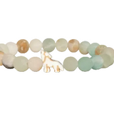 A beaded stretch Fahlo bracelet featuring round, polished beads in soft pastel shades of sage green, cream, and light brown, with a small white wolf charm at the center, shown on a plain white background. Available at Be Charmed