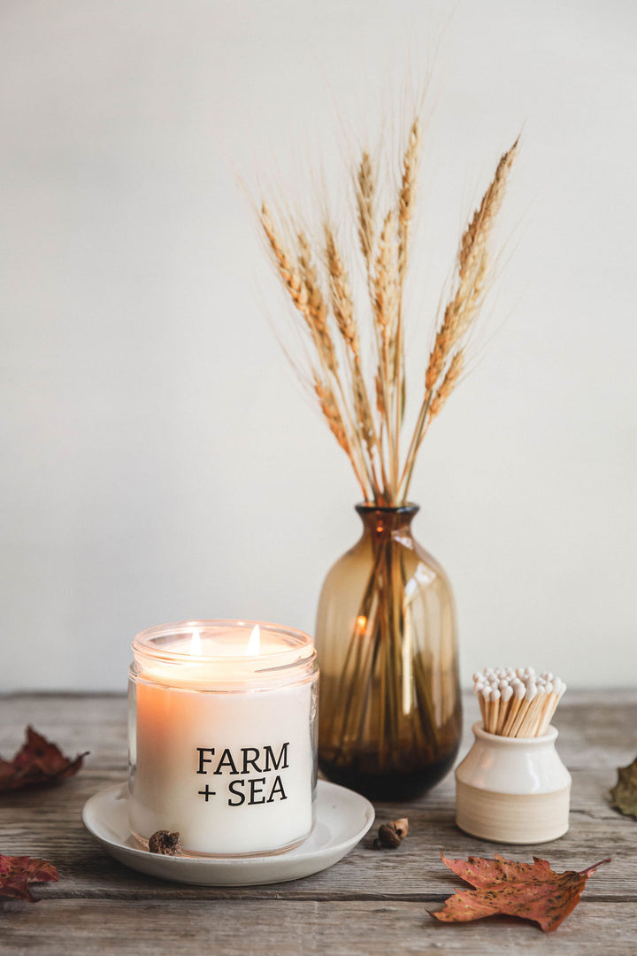 A beige candle with 'Farm + Sea' branding, placed on a wooden surface with a decorative branch and a small white dish with matches. Available at Be Charmed