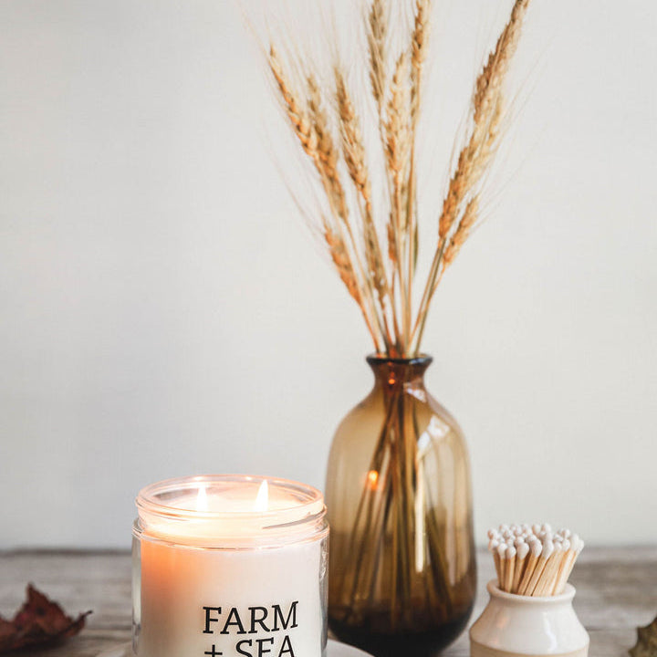 A beige candle with 'Farm + Sea' branding, placed on a wooden surface with a decorative branch and a small white dish with matches. Available at Be Charmed