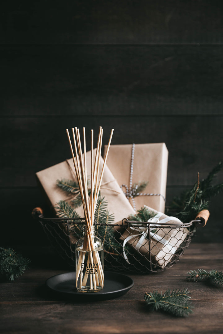 A reed diffuser with rattan reeds in a clear glass bottle, surrounded by a Christmas-themed setup with greenery and gift boxes. Available at Be Charmed