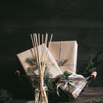 A reed diffuser with rattan reeds in a clear glass bottle, surrounded by a Christmas-themed setup with greenery and gift boxes. Available at Be Charmed