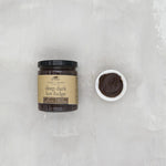 Jar of deep dark hot fudge with a small bowl of fudge on a light gray background. Available at Be Charmed