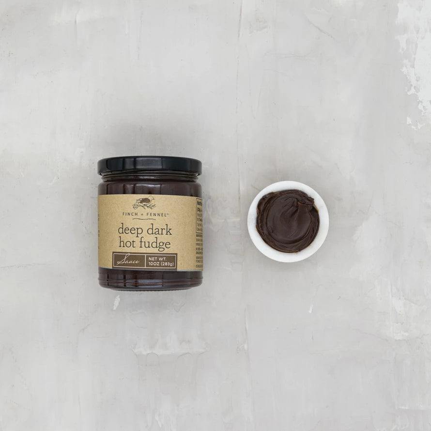 Jar of deep dark hot fudge with a small bowl of fudge on a light gray background. Available at Be Charmed
