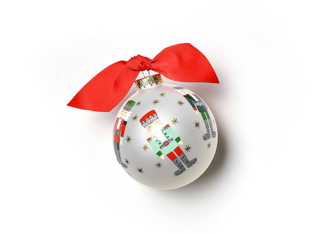 Front view of Coton Colors hand-painted Nutcracker glass ornament with festive gold-star accents, wrapped red ribbon topper, includes matching gift box—Available at Be Charmed