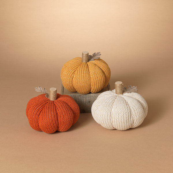 Three knitted pumpkins in orange, yellow, and white on a beige background. Available at Be Charmed.