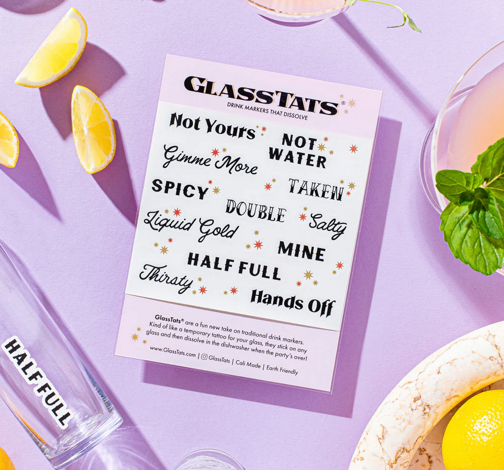 A collection of dissolvable drink markers from Glass Tats laid out on a table with a glass of water and lemon slices. The markers have various playful phrases written on them. Available at Be Charmed